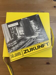 Buchcover "50 Jahre Zukunft" - schwarzweisses futuristisches Gebäude mit gelb hinterlegtem Titel. Das Buch liegt auf einem gelben Skizzenbuch auf einem Holztisch.