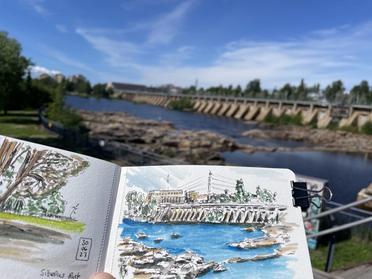 Foto - im Vordergrund ein kleines A6 Aquarellskizzenbuch quer, mit Aquarellzeichnung auf der rechten Seite. Dahinter sichtbar das Motiv - ein Flusssperrwerk mit Überlauf in Oulu